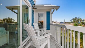 A balcony with white Adirondack chairs, a glass door, and a view of palm trees and houses.