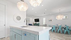 A modern kitchen with a large island, light blue cabinets, and a white marble countertop. There is a dining area with turquoise chairs and a chandelier.