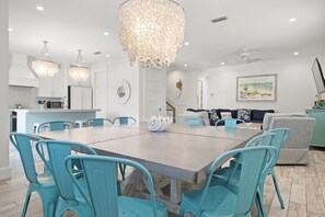 Un vaste espace de salle à manger avec une grande table et des chaises turquoise, une cuisine dotée de meubles blancs et un lustre.