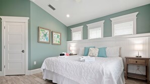 A bedroom with a large bed, white bedding, and two bedside lamps. There are two framed artworks on the wall and a door to the left.