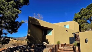 Une maison moderne avec un chemin de pierre menant à l’entrée, entourée d’une végétation aride et sous un ciel bleu dégagé.