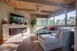 A modern living room with a stone fireplace, a flat-screen TV, leather sofa, and large windows offering a view of greenery.