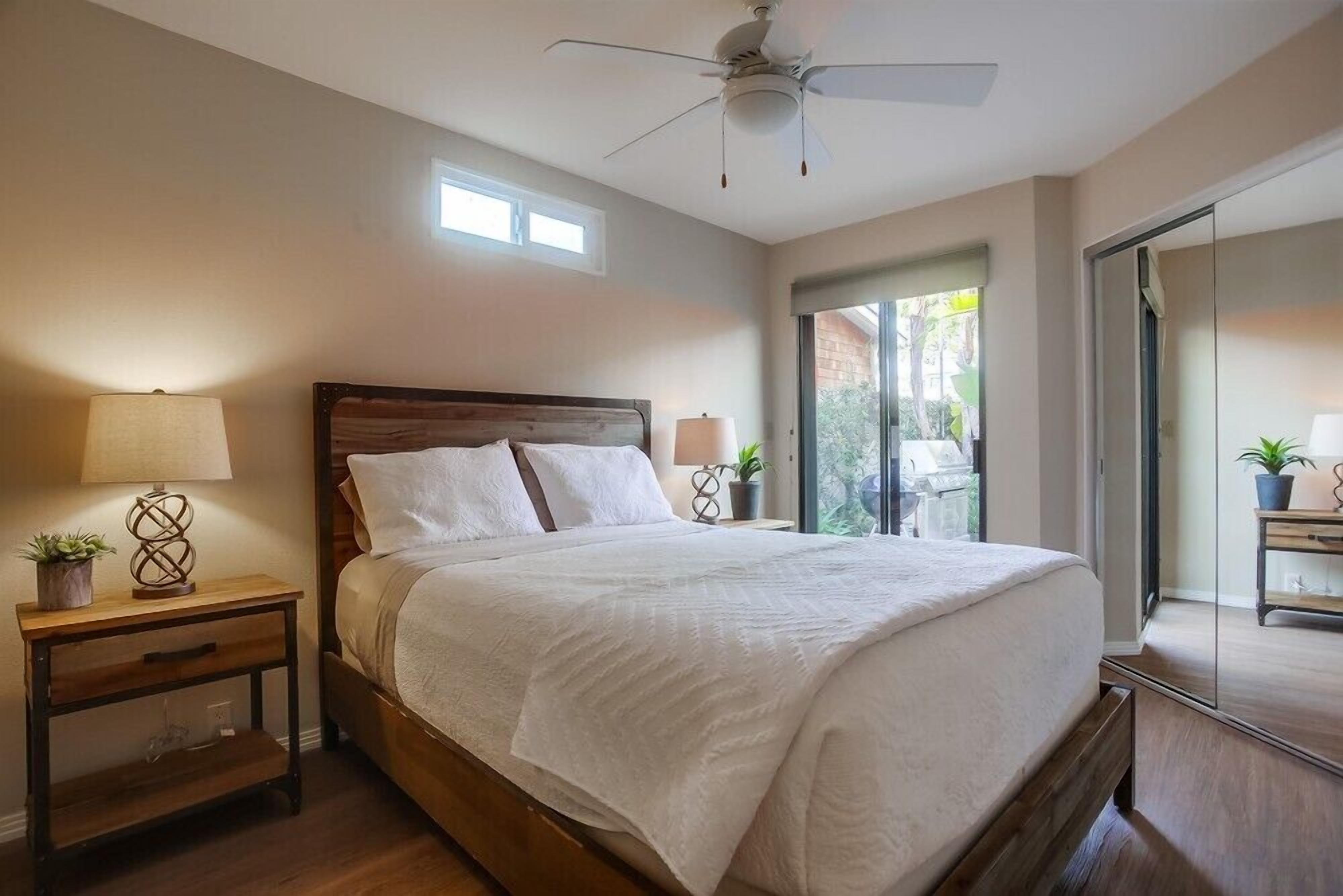 A bedroom with a large bed, wooden headboard, two bedside tables with lamps, a ceiling fan, and a sliding glass door leading to an outdoor area.