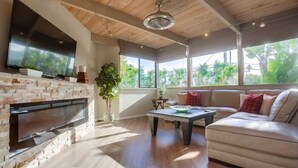 A modern living room with a stone fireplace, a flat-screen TV, a sofa, and large windows offering a view of greenery. - 1.5 Blocks From The Beach: The High Tide  in Del Mar (Del Mar)