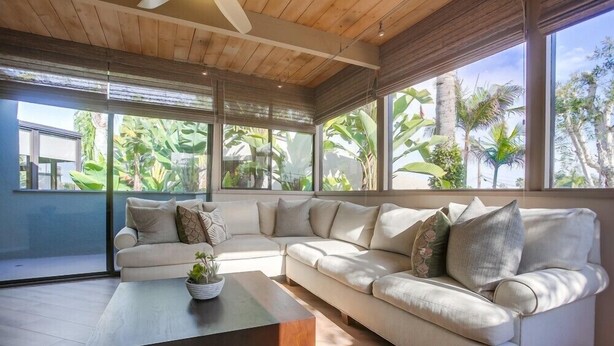 A modern living room with a large sectional sofa, a coffee table, and large windows offering a view of tropical plants.