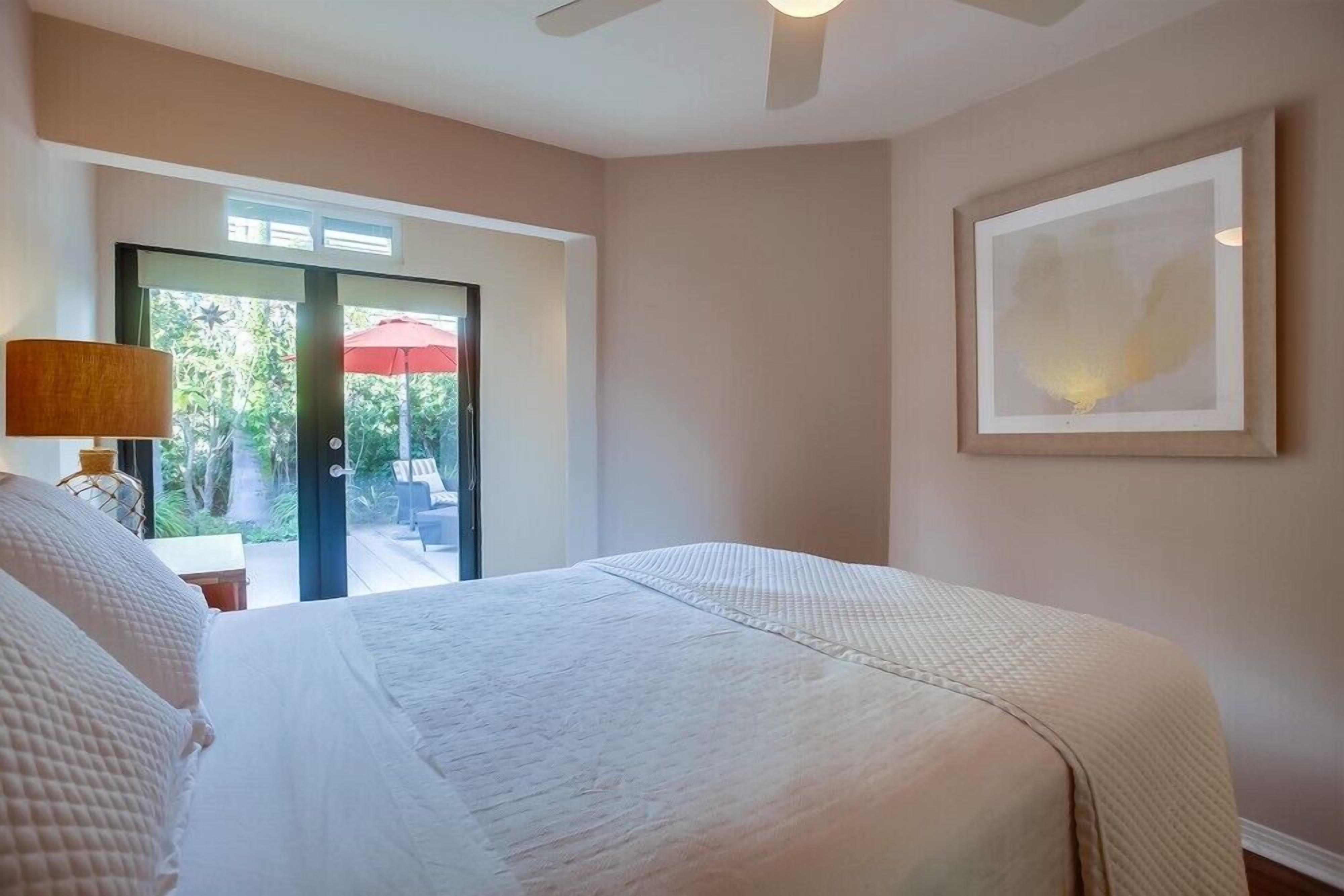 A bedroom with a large bed, a ceiling fan, a window with a view of an outdoor area, and a framed picture on the wall.