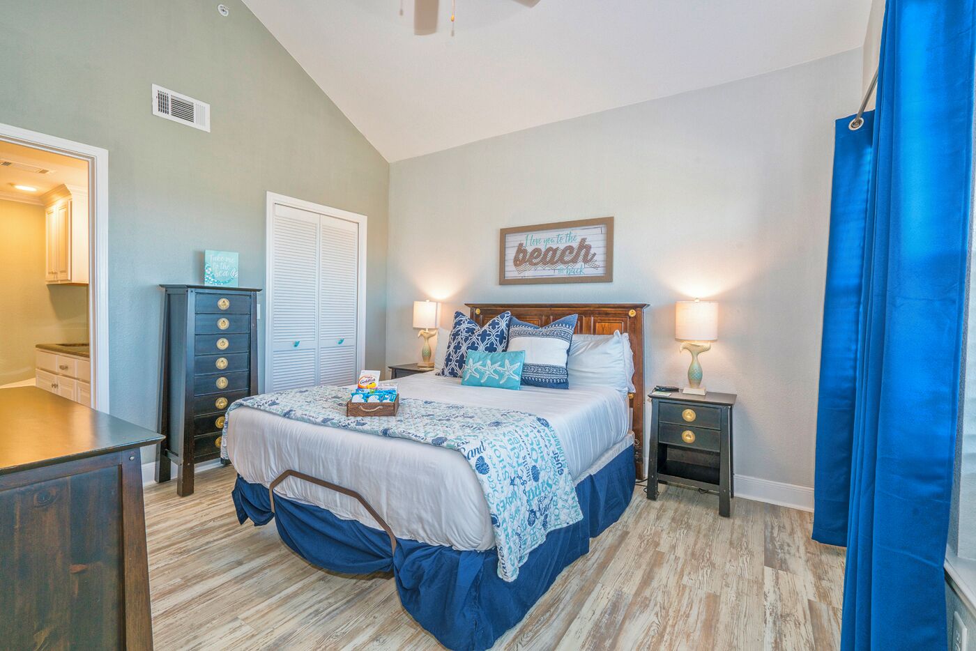 A bedroom with a bed, bedside tables, a dresser, a window with curtains, and a wall sign.