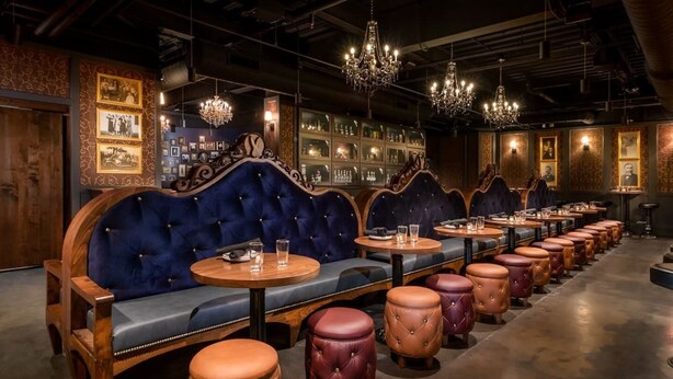 A row of wooden tables with round wooden stools, a blue upholstered bench, and chandeliers in a dimly lit room with framed pictures on the walls. - Awesome Downtown Gilbert location | Pets welcome (Gilbert)