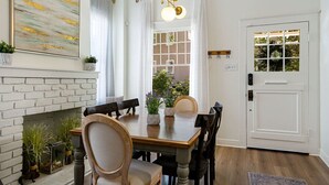 Ein Esszimmer mit Kamin aus Ziegelsteinen, einem Holzesstisch und Stühlen. Eine weiße Tür mit Fenster und eine Hängeleuchte über dem Tisch sind ebenfalls zu sehen.
