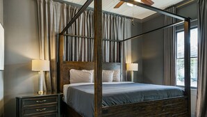 Ein Bett mit Baldachin, ein hölzerner Rahmen und graue Bettwäsche, ein Nachttisch mit Schubladen und ein Deckenventilator.