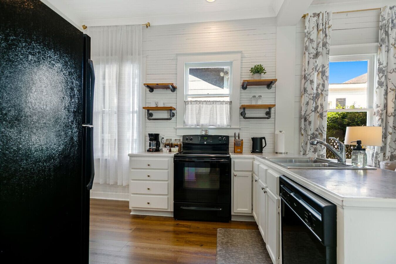 Una cucina moderna con mobili bianchi, un piano cottura nero e una lavastoviglie. Si notano scaffali in legno sopra il piano cottura e una finestra con tende bianche.