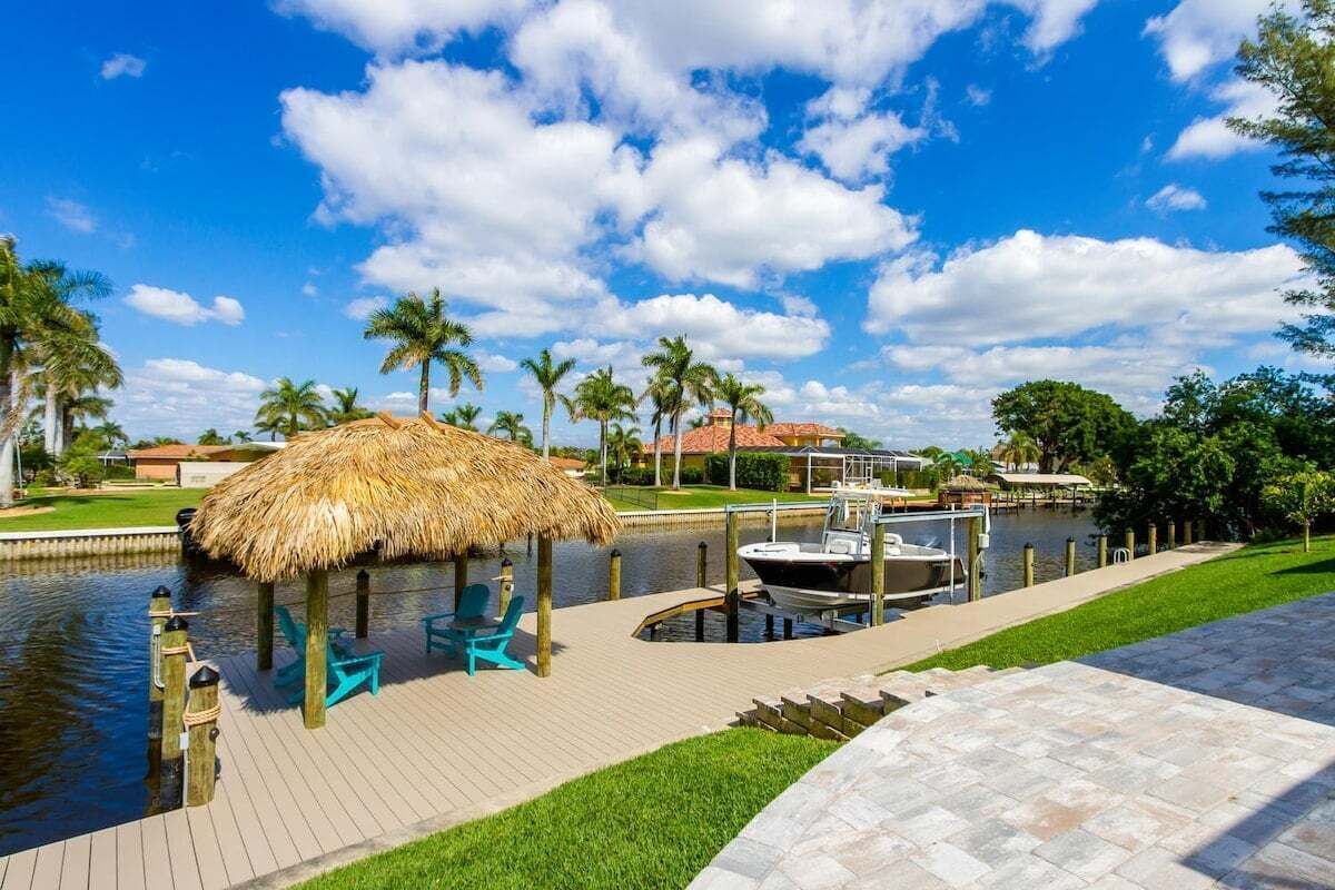 A waterfront area with a thatched-roof structure, a dock with a boat, and a paved walkway.