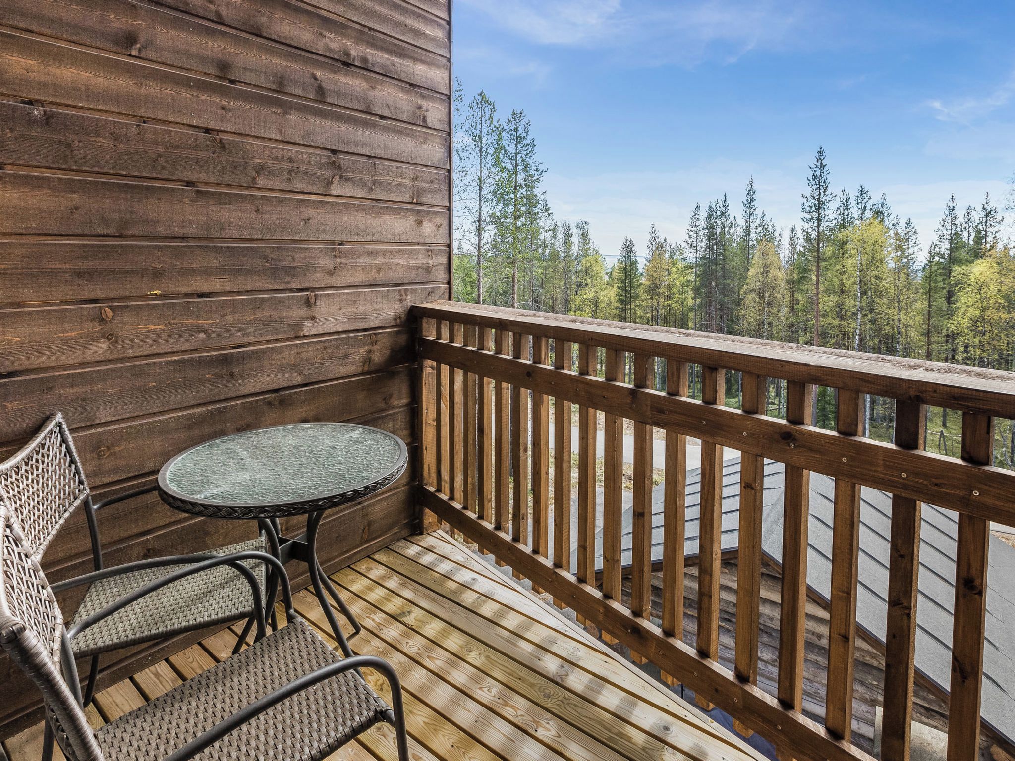 A wooden deck with a round table and two wicker chairs, overlooking a forested area.