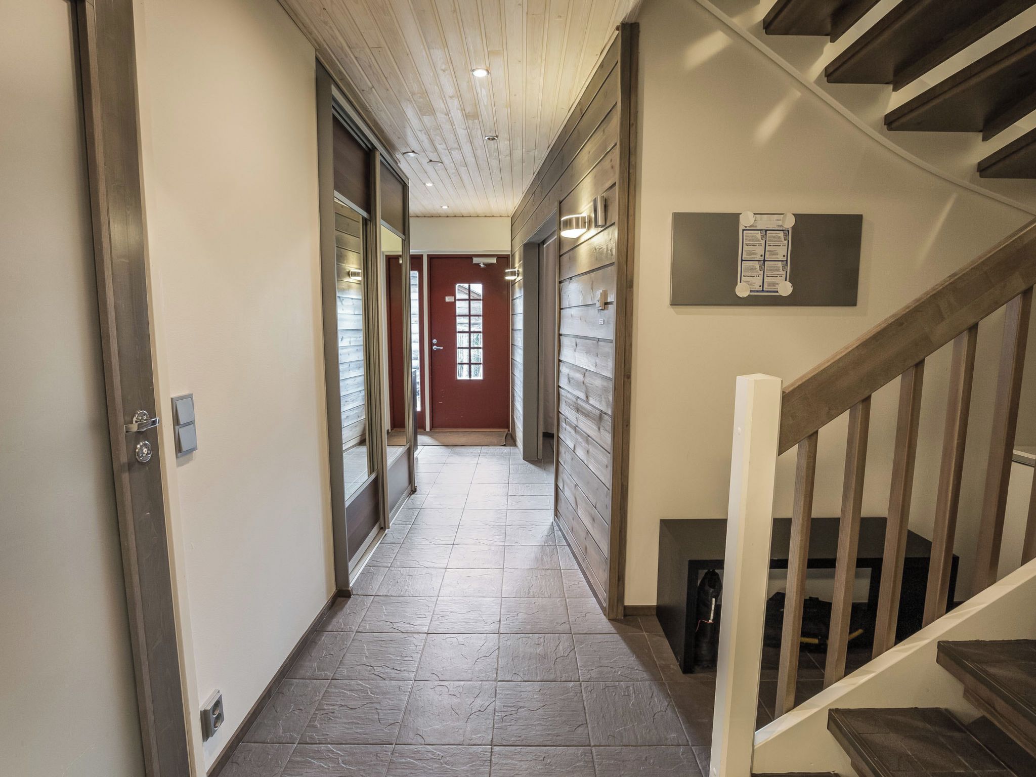 Ein Flur mit einer Holztreppe rechts, einer roten Tür am Ende und einer Pinwand an der Wand.