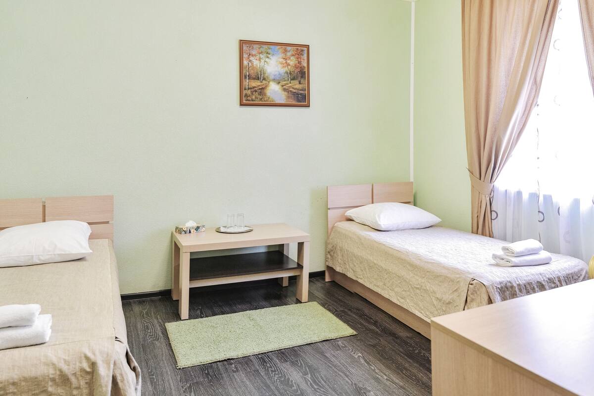 A hotel room with two beds, a small table with a lamp, a painting on the wall, and a window with curtains.