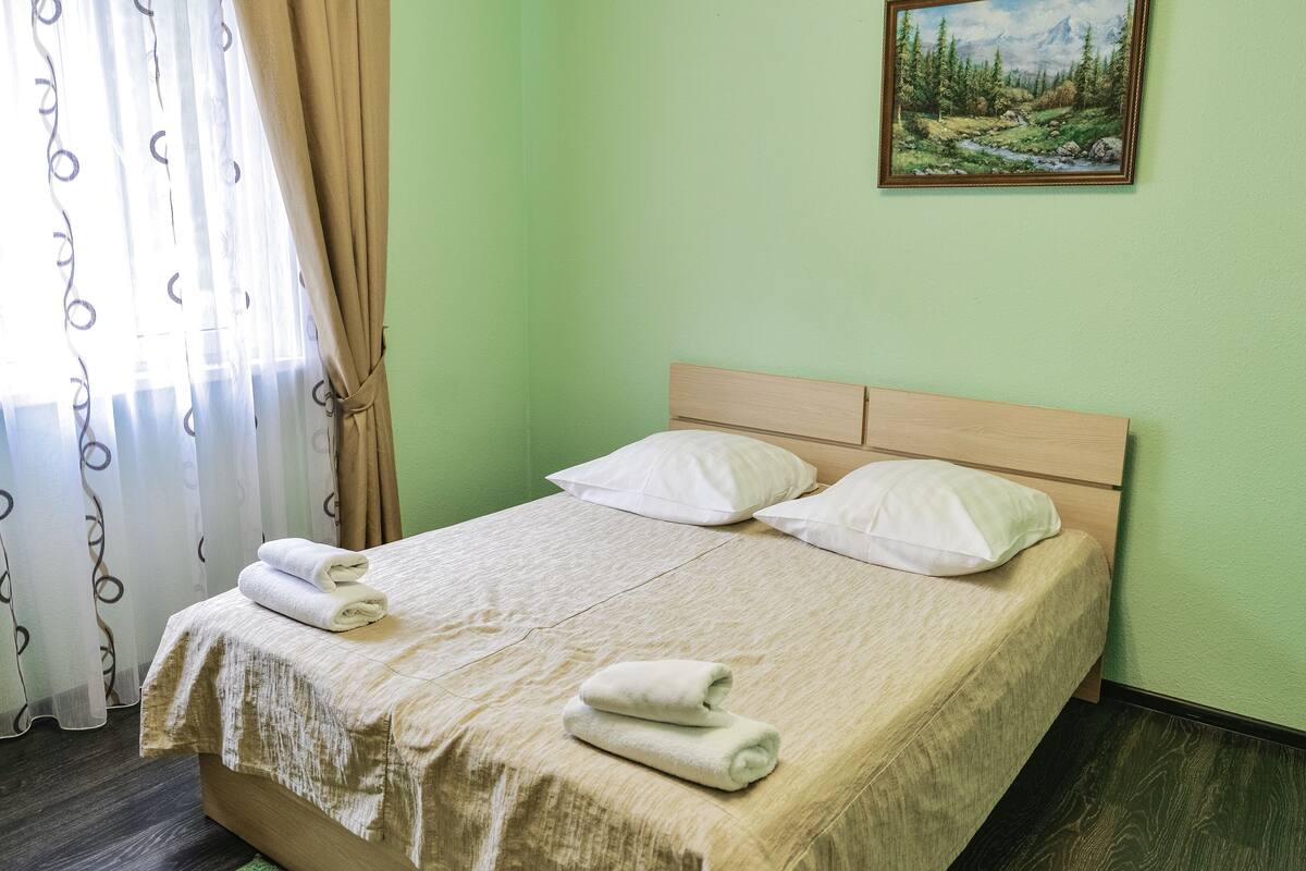 A neatly made bed with two pillows and two folded towels, a painting of a landscape on the wall, and a window with curtains.