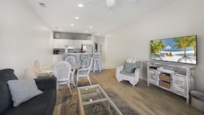 A modern living room with a black sofa, white wicker chairs, a glass coffee table, a TV on a wooden stand, and a kitchen in the background. - Book your Sea Trail getaway in this ground floor, pet friendly condo (Sunset Beach)