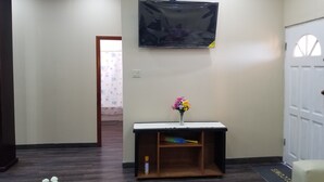 A room with a flat-screen TV mounted on the wall, a small wooden console table with a vase of flowers, and a white door with a window.