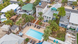An aerial view of a residential area with a swimming pool surrounded by lounge chairs and umbrellas, a fenced yard, and various houses with different architectural styles.