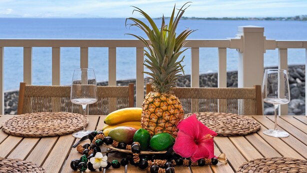 Uma mesa de madeira com um jogo americano trançado, uma abacaxi, bananas, limas, uma flor de hibisco e um copo de vinho branco.