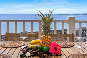 Una mesa de madera con un mantel tejido, una piña, plátanos, limas, una flor de hibisco y una copa de vino blanco.