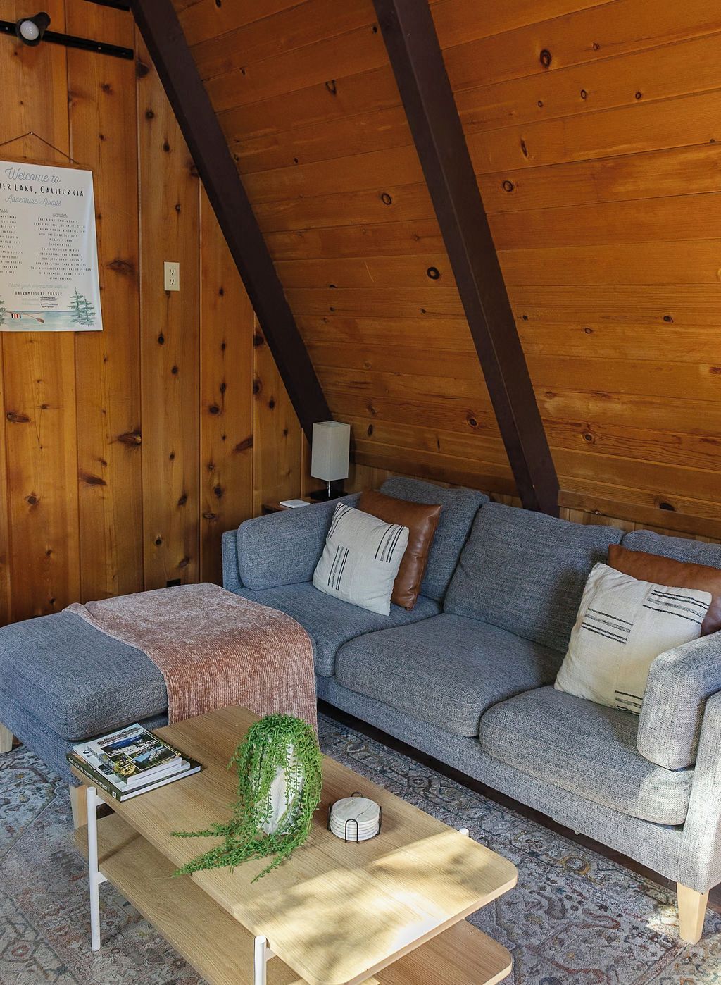 Un espacio acogedor con techo de madera, un sofá gris con cojines estampados, una mesita con una planta y un libro, y un cartel de bienvenida en la pared.