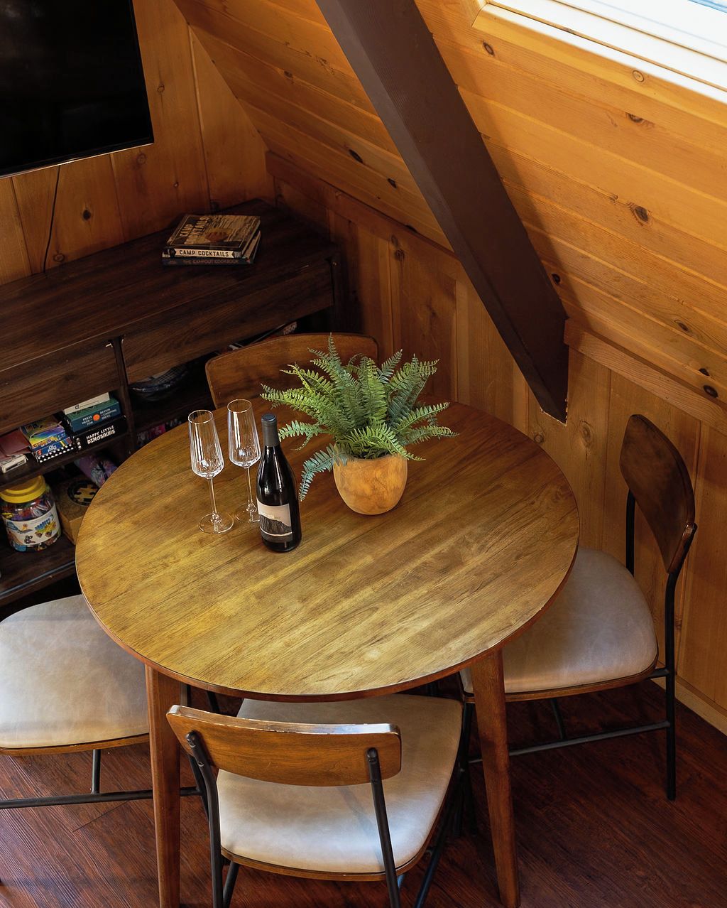 A wooden interior with a round table, two chairs, a bottle of wine, two glasses, and a potted plant.