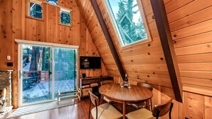 A cozy wooden cabin with a dining area, a fireplace, and large windows offering views of the forest.