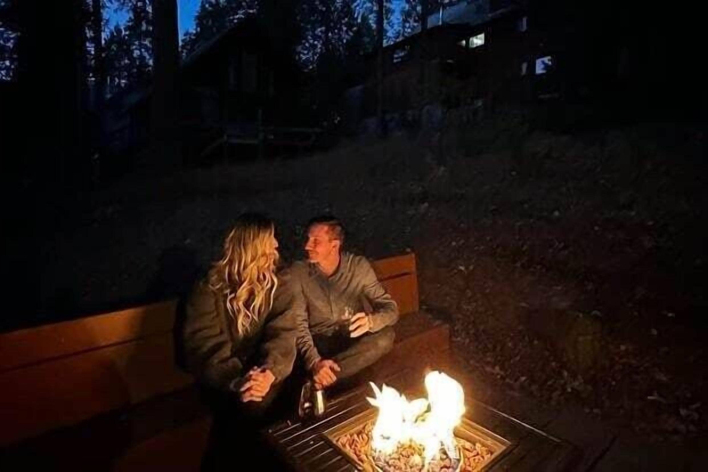 Two people sitting on a bench outdoors at night, with a campfire in front of them.