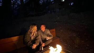 Two people sitting on a bench outdoors at night, with a campfire in front of them.