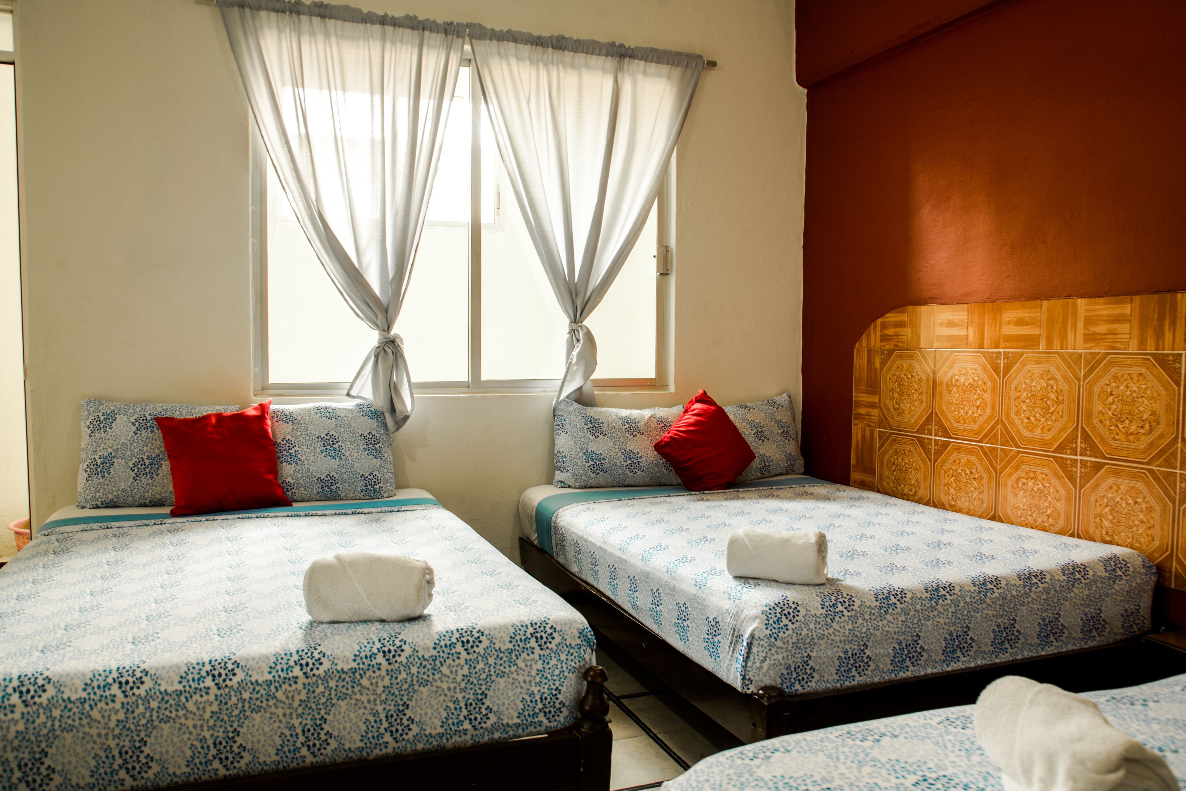 Habitación de hotel con dos camas, cada una con una colcha estampada y almohadas rojas. Se ve una ventana con cortinas de gasa, y las paredes tienen un papel tapiz estampado.