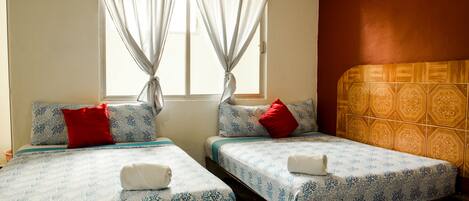 Habitación de hotel con dos camas, cada una con una colcha estampada y almohadas rojas. Se ve una ventana con cortinas de gasa, y las paredes tienen un papel tapiz estampado.
