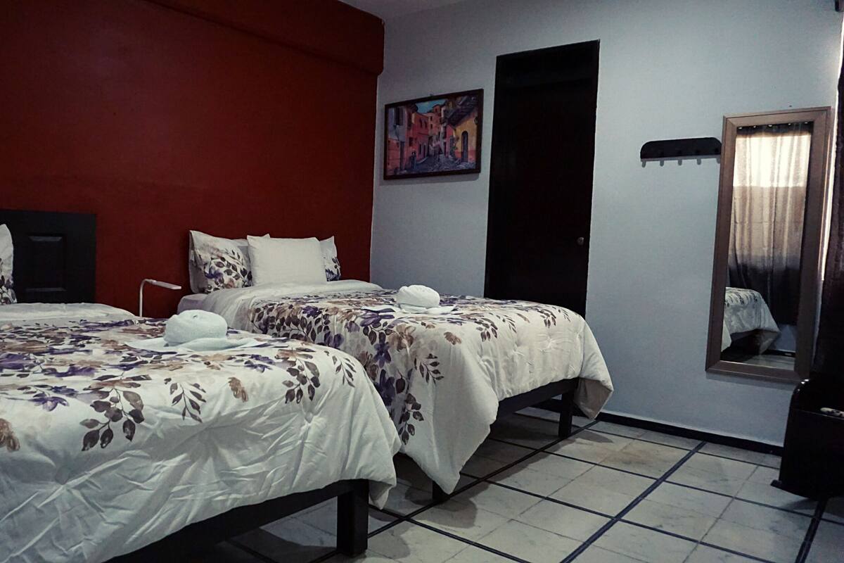 A hotel room with two beds, a red accent wall, a painting, and a mirror.