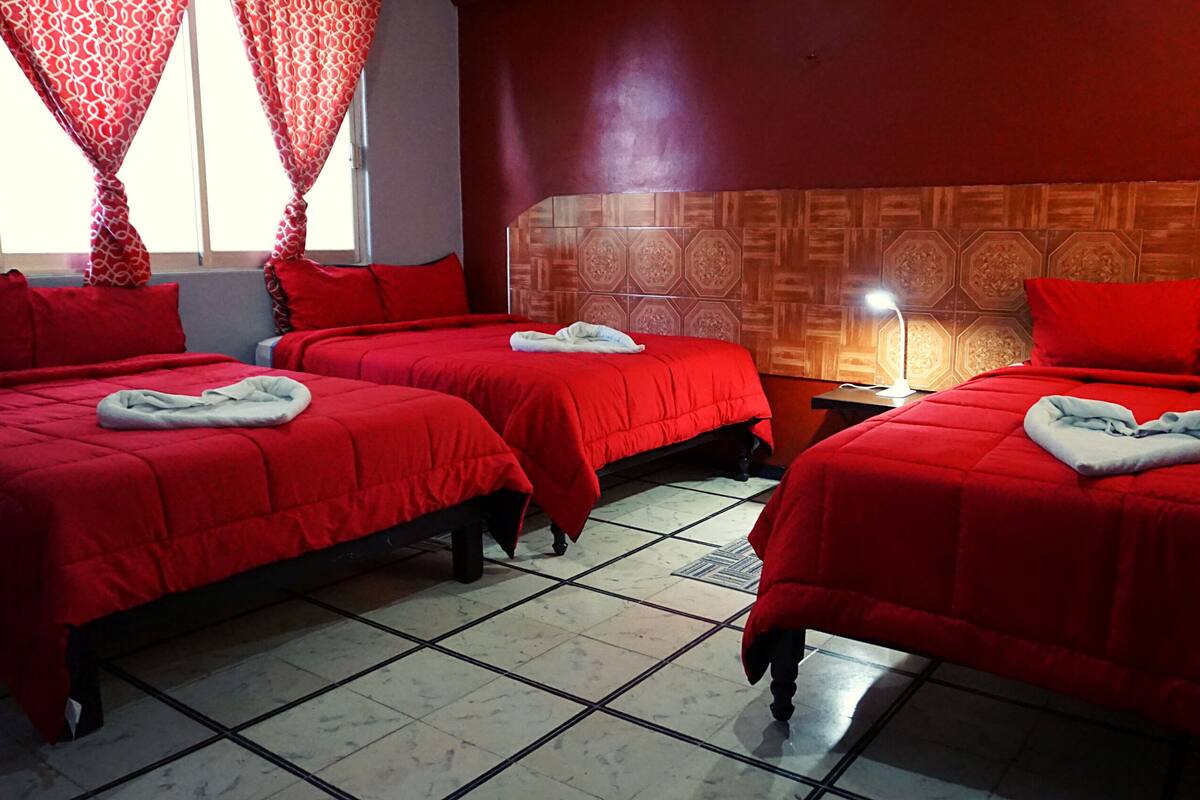 A hotel room with two beds, red bedding, a patterned headboard, a nightstand with a lamp, and a window with red curtains.