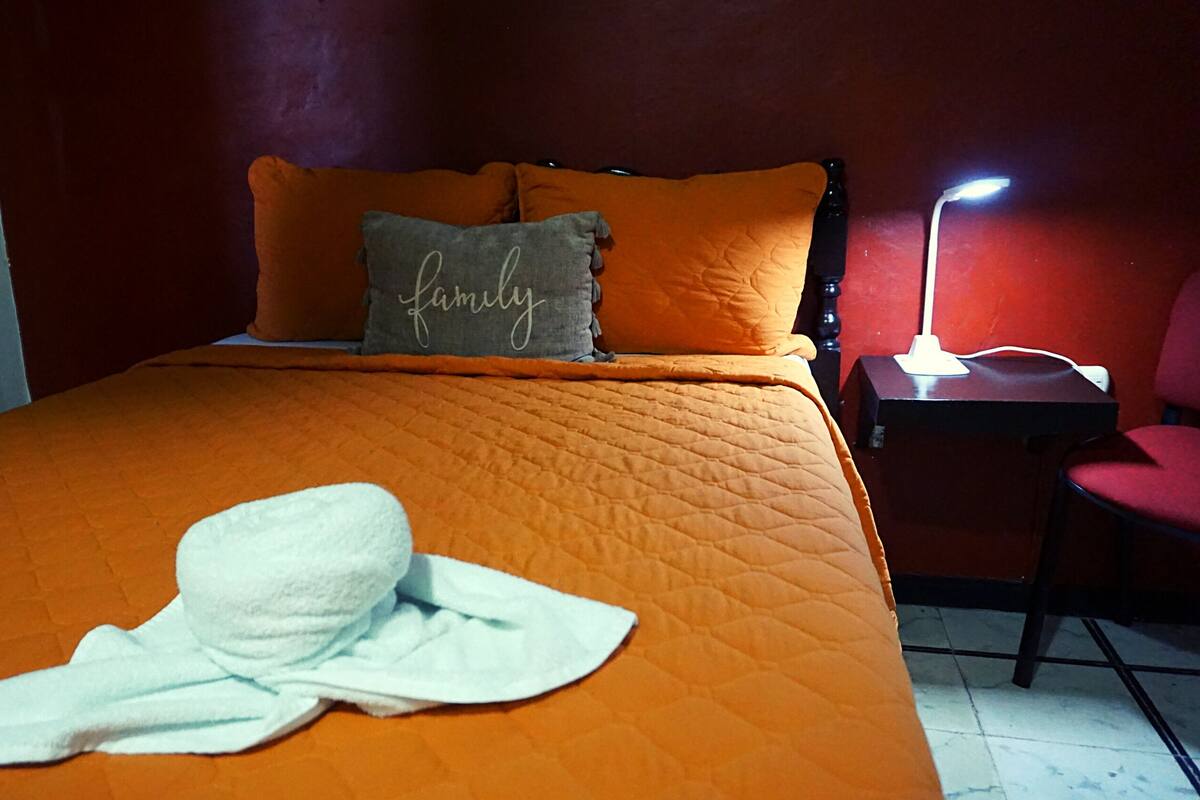 A bed with an orange quilt, a gray pillow with the word "family" on it, and a white towel on the bed.