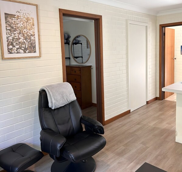 Une pièce avec un fauteuil en cuir noir, une commode en bois, un mur en briques blanches et une porte donnant sur une autre pièce.