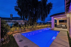 Ein Schwimmbecken mit blau beleuchtetem Rand, umgeben von einem Zaun und Bäumen, mit einem überdachten Sitzbereich rechts.