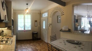 A kitchen with a gas stove, a tiled floor, a dining table with a white tablecloth, and a window with curtains. - VILLA 230 m² - 16 Pers - 8chambres -4000m² de parc /piscine 10 x 5m a 5 mn plage (Argelès-sur-Mer)