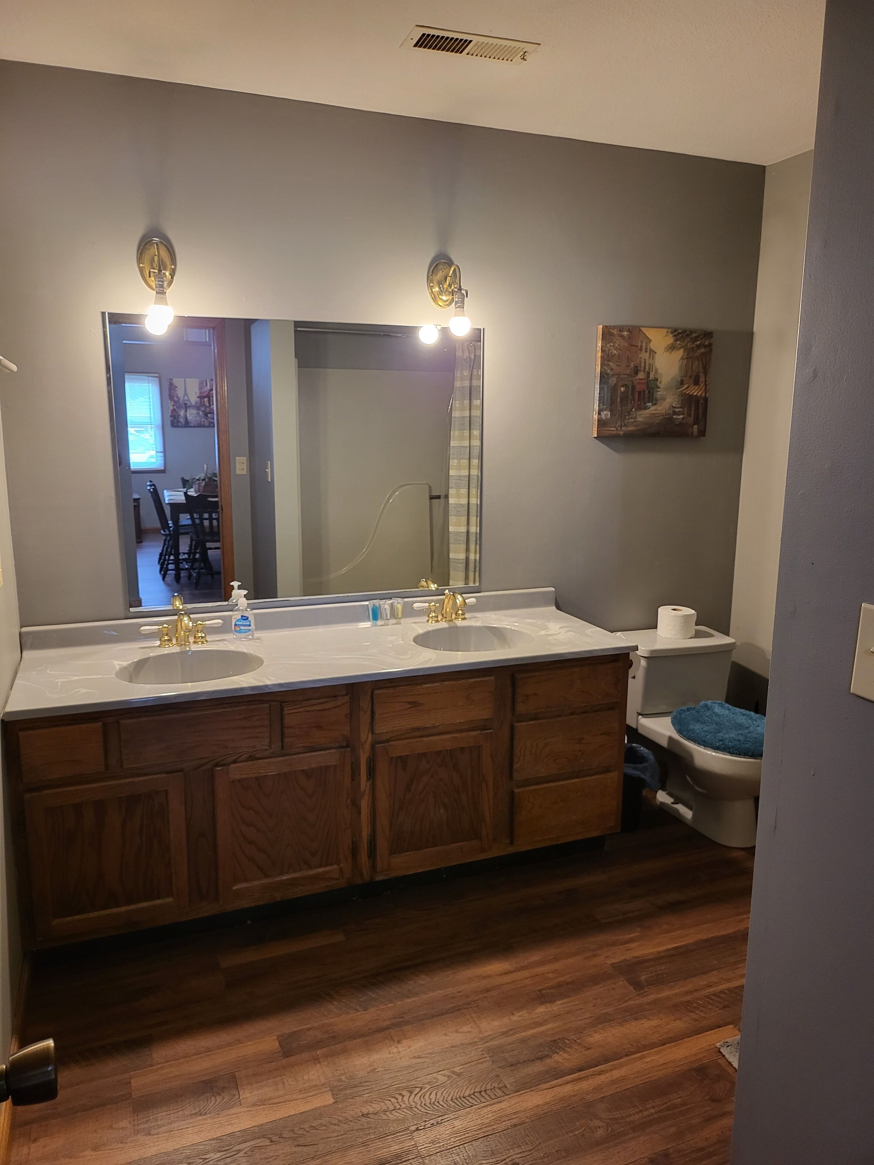 A bathroom with a double sink vanity, wooden cabinets, a large mirror with lights, a toilet with a blue bath mat, and a painting on the wall.