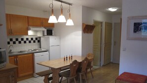 Cocina compacta con armarios de madera, una estufa blanca y una mesa de comedor con sillas. Hay un pequeño banco con cojín rojo y un cuadro en la pared.