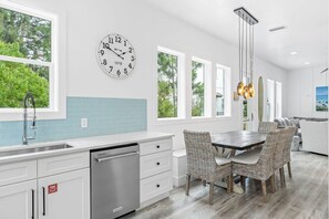 Une cuisine moderne avec une horloge, des meubles blancs et un coin repas avec des chaises en osier, offrant une vue sur la verdure à l’extérieur.