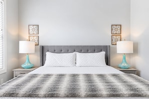A bedroom with a tufted headboard, a fluffy gray blanket, two bedside tables with decorative lamps, and wall art.