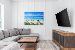 En stue med en grå sofa, et sofabord, et fladskærms-tv og et maleri af en strand.