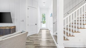 A hallway with a wooden floor, white walls, a TV on a wooden cabinet, and a staircase with white railings. - "Shore To Please" - Cottages at Seagrove - 5 Bed / 5.5 Bath - Sleeps 14 - Across from the Pool! (Santa Rosa Beach)