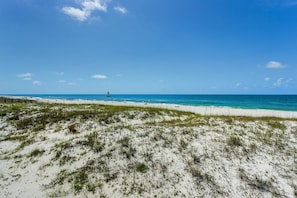 A beach with white sand and sparse vegetation, a clear blue sky, and a distant sailboat. - Beautiful Villa in Lost Key | EV Charger | Screened Balcony | Private Beach Club | Pools | Free Golf (Pensacola)
