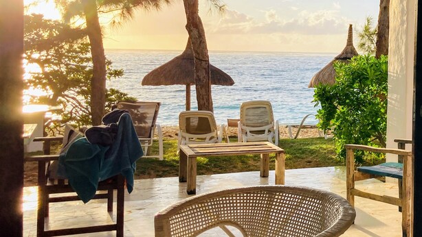 Een terras aan het strand met loungestoelen, een tafel en uitzicht op de oceaan bij zonsondergang.
