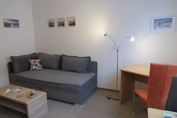 A room with a grey sofa, a wooden desk, a red chair, a floor lamp, and a small table with a book and a bowl.