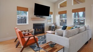 A modern living room with a fireplace, a flat-screen TV, and a large window with a view of trees. - Dwell 30 Day Minimum- Modern & Peaceful, Walk to Old Mill, New and Lovely, AC (Bend)