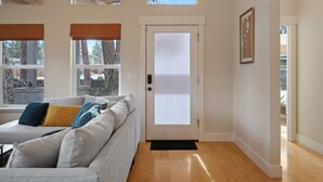 A modern living room with a grey sofa, a door with frosted glass, and large windows with blinds. - Dwell 30 Day Minimum- Modern & Peaceful, Walk to Old Mill, New and Lovely, AC (Bend)