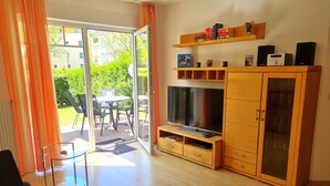 A modern living room with a TV, wooden cabinets, and a glass door leading to an outdoor area. - Vacation apartment 25 Nachtigallenweg (Graal-Müritz)